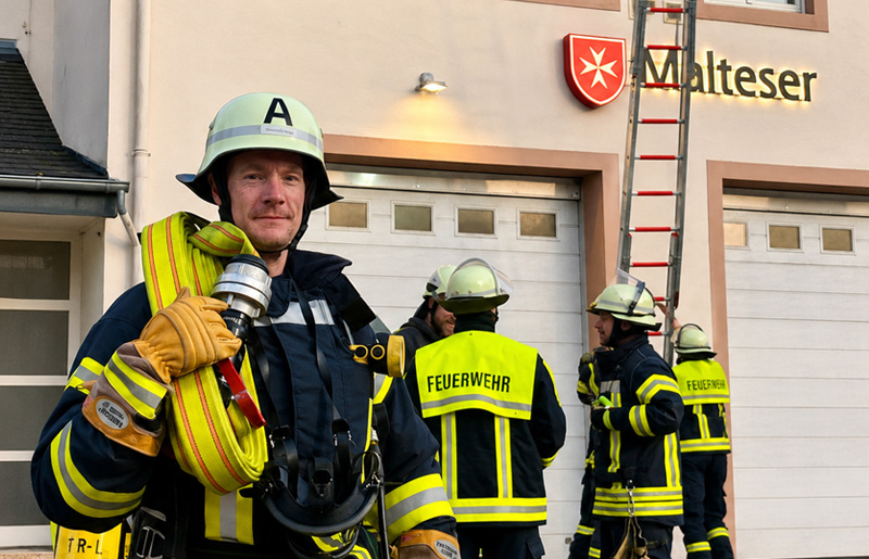 Christoph Biegel steht in Feuerwehrschutzkleidung mit einem Wasserschlauch auf den Schultern vor einem Malteser-Gebäude. Im Hintergrund sind weitere Feuerwehrmänner zu sehen.