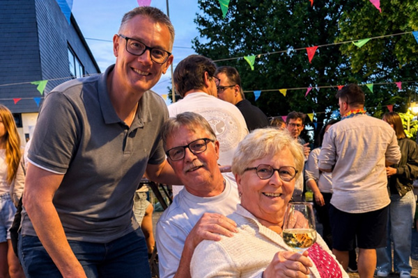 Christoph Biegel beugt sich zu einem sitzenden Mann und einer sitzenden Frau; die Frau präsentiert ein Glas Wein, die Gruppe lächelt freundlich in die Kamera.
